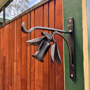 May include: A decorative, black metal wall-mounted hook in the shape of a flower. The hook has a detailed floral design with leaves and a bell-shaped bloom. It is attached to a rectangular plate. The background shows a wooden fence.