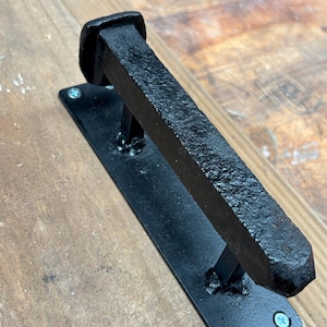 May include: Black metal door handle with a square, flat base and a long, rectangular, textured metal bar. The handle is attached to a wooden surface with screws.