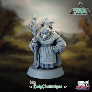 Puede incluir: Una figura gris de una bruja, Lady Cladderdyne, sosteniendo un bastón con un cristal. Lleva una túnica con capucha y un delantal. La base es cuadrada. Los textos "Warriors & Warrens" y "Broken Anvil Monthly" son visibles.