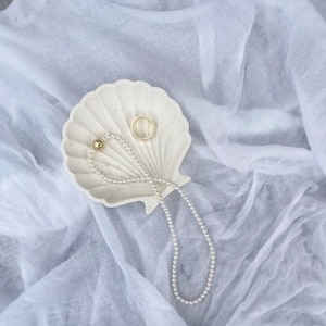 Può includere: Un piatto in ceramica bianca a forma di conchiglia con una collana di perle e un anello d'oro su uno sfondo di tessuto bianco.