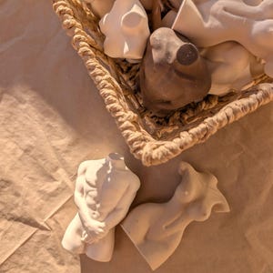 May include: A collection of small, white and brown torso sculptures in a woven basket. The sculptures are of male and female forms, with detailed musculature and curves. The basket sits on a light brown surface.