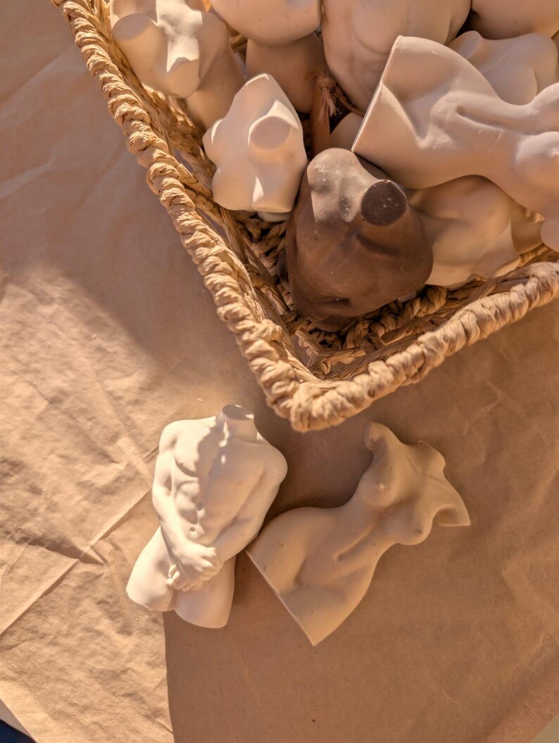 May include: A collection of small, white and brown torso sculptures in a woven basket. The sculptures are of male and female forms, with detailed musculature and curves. The basket sits on a light brown surface.