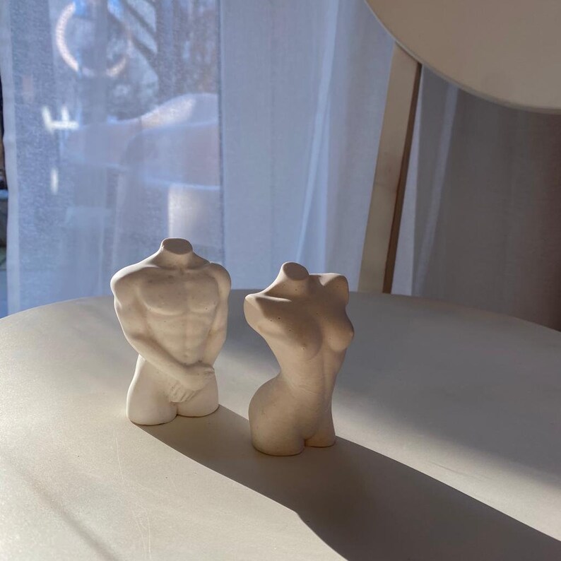 May include: Two off-white, headless torso sculptures, one male and one female, stand on a white surface. The male figure has defined musculature, while the female figure has a curved form. The sculptures are lit by natural light.