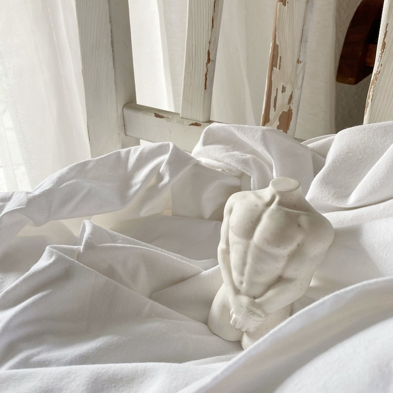 May include: A white, headless torso sculpture with defined musculature, resting on a bed of white fabric. The sculpture's arms are crossed in front. The background includes a white chair and sheer curtains.