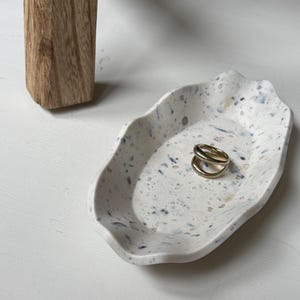 Può includere: Un vassoio decorativo maculato bianco sporco con un bordo ondulato, che contiene un anello d'oro. Il vassoio ha macchie blu e grigie. Una gamba di tavolo in legno è visibile sullo sfondo.