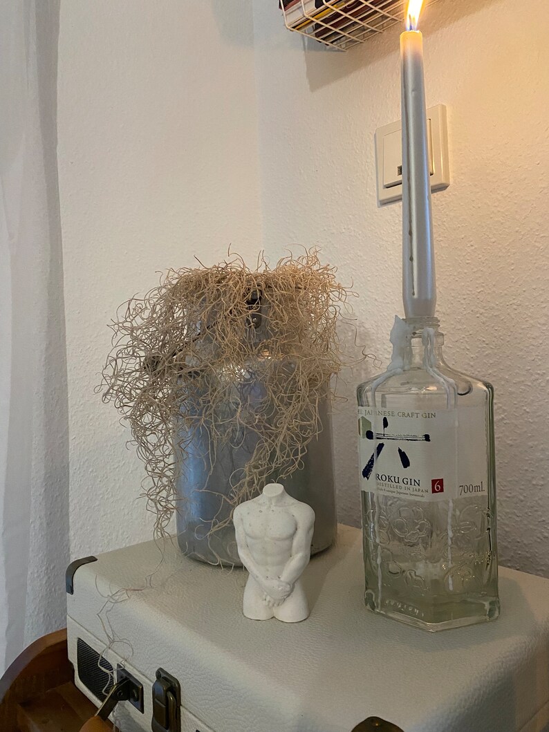 May include: A decorative arrangement featuring a white candle burning atop a clear gin bottle, a gray vase with dried plant material, and a small white torso statue. The bottle has a label that reads "ROKU GIN". The items are on a white surface.
