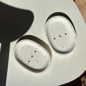 Op de afbeelding: Twee gevlekte, ovale zeepbakjes. De bakjes zijn gebroken wit met kleine, donkerbruine spikkels. Elk bakje heeft drie afvoergaten. De items worden gepresenteerd op een wit oppervlak, met een donkere schaduw op de achtergrond.