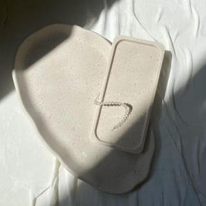 Op de afbeelding: Een lichtbeige, gespikkelde, hartvormige dienblad met een bijpassend rechthoekig dienblad. Een heldere kralenarmband ligt op het rechthoekige dienblad. De items staan op een witte, getextureerde ondergrond, met schaduwen van zonlicht.