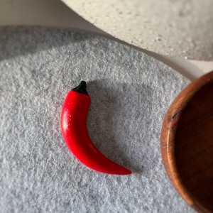 Può includere: Una vivace statuetta di peperoncino rosso con finitura lucida e gambo nero. Il peperoncino è posto su una superficie di feltro grigio, con una ciotola di legno e un oggetto bianco sullo sfondo. Il peperoncino è un oggetto decorativo.