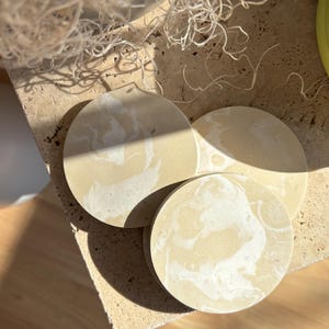 Könnte beinhalten: Drei runde beige Untersetzer mit einem weißen Marmormuster. Die Untersetzer befinden sich auf einer hellbraunen Oberfläche.