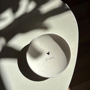 Könnte beinhalten: Eine kleine, cremefarbene Keramikschale mit einem schwarzen Herz und dem Wort "frieda". Die Schale hat eine gesprenkelte Textur und steht auf einer hellen Oberfläche. Das Bild wird von Sonnenlicht beleuchtet, das Schatten wirft.