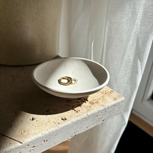 Könnte beinhalten: Eine kleine, cremefarbene Keramikschale enthält einen goldenen Ring und einen kleinen Perlenring. Die Schale steht auf einer strukturierten, hellen Oberfläche, mit einem weichen weißen Vorhang im Hintergrund. Die Szene wird von natürlichem Licht beleuchtet.