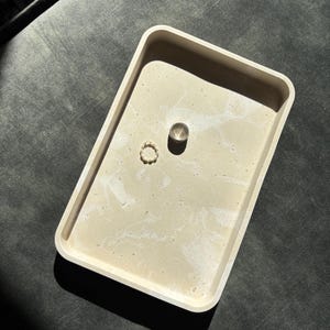 Op de afbeelding: Een rechthoekig beige dienblad met een gemarmerd wit patroon. Twee ringen, een zilveren en een gouden, liggen in het dienblad. Het dienblad staat op een donkergrijze ondergrond.