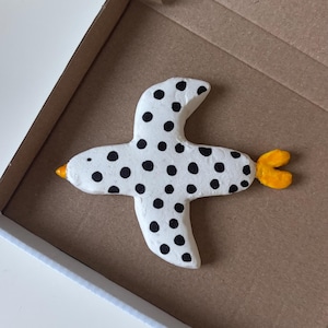 Könnte beinhalten: Eine weiße Vogelfigur mit schwarzen Punkten, einem kleinen orangefarbenen Schnabel und einem leuchtend orangefarbenen Schwanz. Der Vogel ist mit ausgebreiteten Flügeln geformt und liegt in einer einfachen, offenen braunen Pappschachtel.