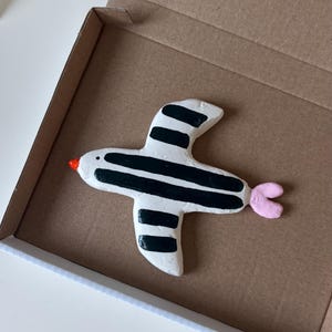 Könnte beinhalten: Eine handgeformte, weiße Vogelfigur mit markanten schwarzen vertikalen Streifen auf den Flügeln und dem Körper. Der Vogel hat einen kleinen, leuchtend orangefarbenen Schnabel und einen hellrosa, dreizackigen Schwanz. Er liegt in einer braunen Pappschachtel.