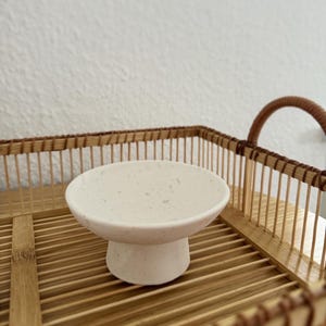 Może przedstawiać: Mała, kremowa ceramiczna miska na nóżce z nakrapianymi detalami. Misa stoi na okrągłej podstawie i jest prezentowana na bambusowej tacy z plecioną krawędzią i uchwytem. Idealna do serwowania małych porcji.