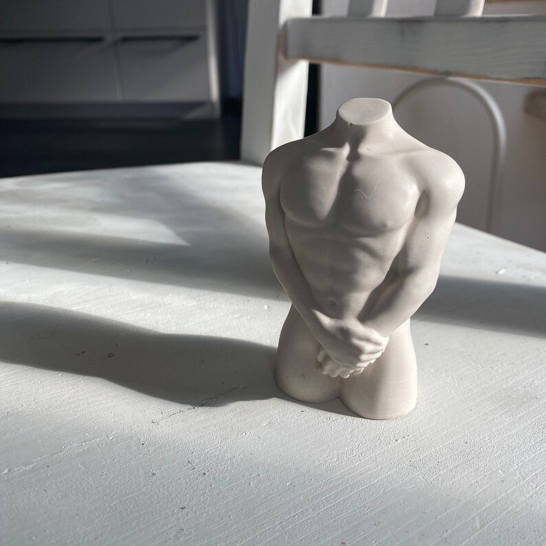 May include: A small, off-white sculpture of a male torso. The figure has a muscular build, with arms crossed over the lower abdomen. The head is missing. The sculpture is on a white surface.