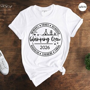 A white t-shirt with a graphic design, a pair of white sneakers, and a small green plant on a wooden surface.