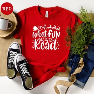 May include: Red t-shirt with white text that reads 'Oh what fun it is to read' with a small image of a Santa in a sleigh with reindeer above the text. The t-shirt is laying on a brown surface with a pair of black sneakers, a pair of blue jeans, and a sprig of green foliage.