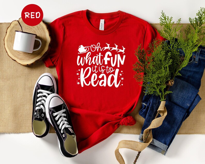May include: Red t-shirt with white text that reads 'Oh what fun it is to read' with a small image of a Santa in a sleigh with reindeer above the text. The t-shirt is laying on a brown surface with a pair of black sneakers, a pair of blue jeans, and a sprig of green foliage.