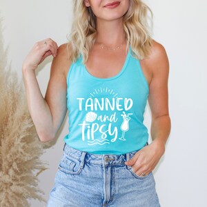 May include: A light blue tank top with the text "Tanned and Tipsy" in white. The text is surrounded by a seashell and a cocktail glass graphic.