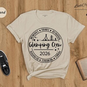 a beige t-shirt with a graphic design, a pair of black sneakers, an open book, and a pair of sandals, all arranged on a wooden surface.