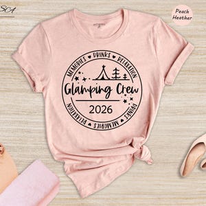 a pink t-shirt with a graphic design, a pair of sunglasses, and a hat, all placed on a wooden surface.