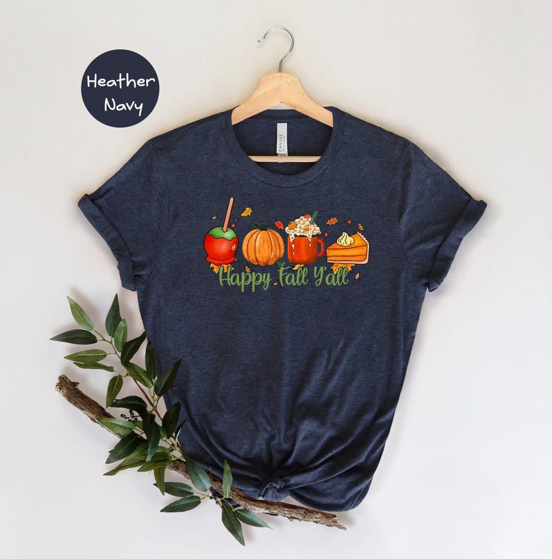 May include: A heather navy blue t-shirt with a graphic of a caramel apple, pumpkin, a mug of hot chocolate with whipped cream, and a slice of pumpkin pie. The text 'Happy Fall Y'all' is printed below the graphic.