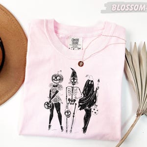 May include: A light pink Comfort Colors t-shirt featuring a Halloween-themed illustration. The graphic depicts three figures: a pumpkin-headed character, a skeleton, and a ghost, each holding a trick-or-treat bag. A gold necklace with a circular pendant is draped on the shirt.