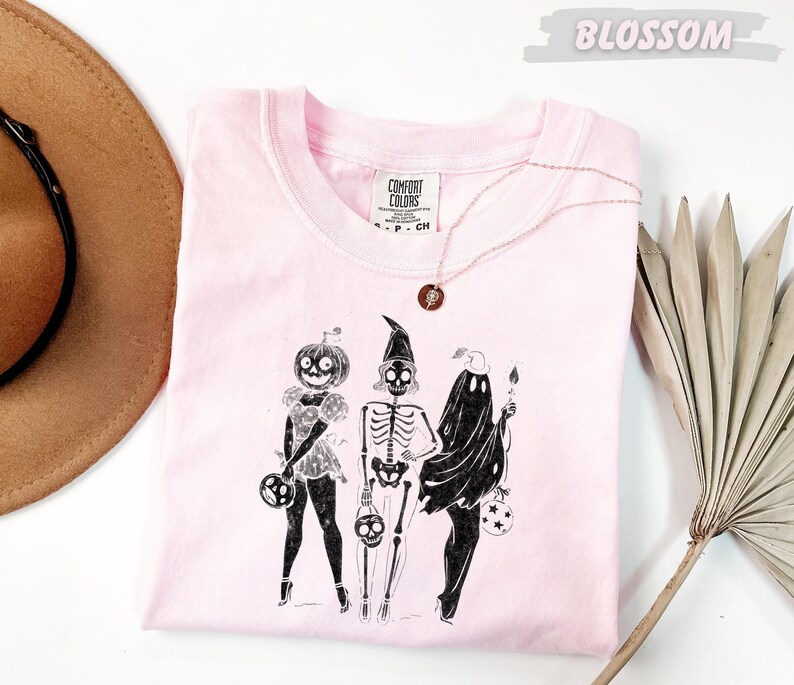 May include: A light pink Comfort Colors t-shirt featuring a Halloween-themed illustration. The graphic depicts three figures: a pumpkin-headed character, a skeleton, and a ghost, each holding a trick-or-treat bag. A gold necklace with a circular pendant is draped on the shirt.