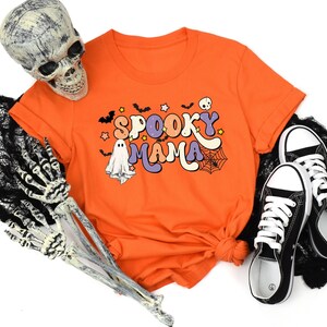 May include: Orange t-shirt with 'Spooky Mama' text in retro style lettering.  The design includes ghosts, bats, stars, and a spiderweb.  Black canvas shoes are also shown in the image.