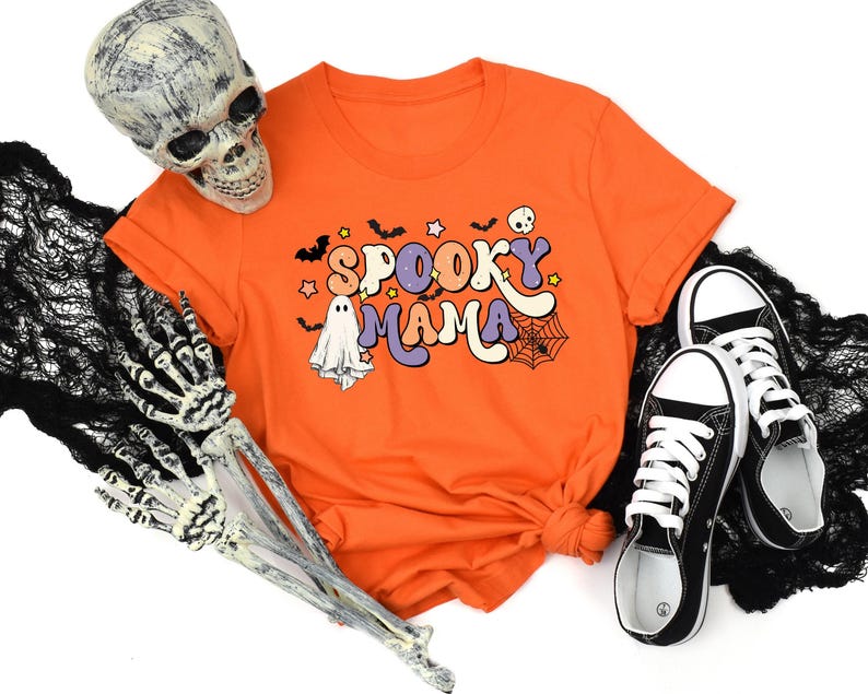 May include: Orange t-shirt with 'Spooky Mama' text in retro style lettering.  The design includes ghosts, bats, stars, and a spiderweb.  Black canvas shoes are also shown in the image.