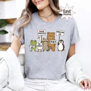 May include: A gray t-shirt featuring a cartoon illustration of a frog, duck, raccoon, bear, and penguin holding signs with various messages. The signs read "F*ck ICE", "No Kings", "I Due Process", "FDT", and "Abolish ICE".