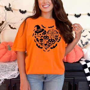 May include: Orange short sleeve t-shirt featuring a black heart-shaped design. The heart is filled with various Halloween icons such as pumpkins, bats, spiders, skulls, and a witch's hat.  The shirt is a comfortable, casual style, perfect for Halloween parties or events.  The design is simple yet festive, making it a great addition to any Halloween wardrobe.