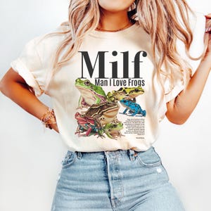 May include: Cream-colored t-shirt featuring a graphic of several colorful frogs and the text "Milf Man I Love Frogs". The frogs are in shades of green, blue, red, and brown. The shirt is made of a soft material.