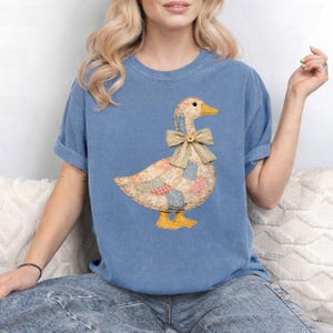 May include: A blue t-shirt featuring a patchwork goose design. The goose has a yellow beak and feet, a bow tie, and a button. The patchwork design incorporates various pastel colors and patterns. The shirt has rolled-up sleeves.