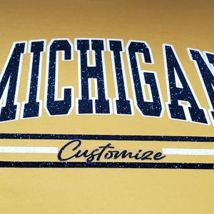 May include: Mustard yellow t-shirt with the word "MICHIGAN" in navy blue, glitter-filled, arched lettering. Below the word "MICHIGAN" is the word "Customize" in a cursive font, with two black stripes and a white stripe.