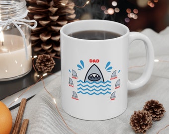 May include: White ceramic coffee mug with a blue and white shark graphic. The text "DAD" is above the shark and the names "MARY", "RUTH", "ESTHER", "ABEL", "JOSEPH", and "DAVID" are around the shark.
