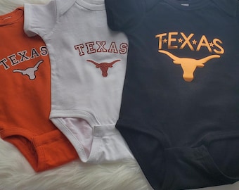 May include: Three baby onesies with the Texas Longhorns logo and the word "TEXAS" printed on them. The onesies are orange, white, and black.