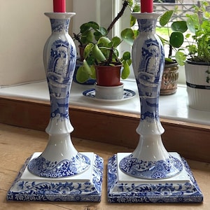 May include: Two blue and white ceramic candle holders with red candles. The candle holders have a floral design and are on a wooden table.