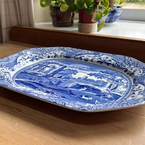 Puede incluir: Una bandeja de servicio rectangular de cerámica con un patrón de transferencia en azul y blanco. La escena central representa un paisaje con ruinas arquitectónicas, árboles y figuras, rodeado por un borde decorativo floral y geométrico.