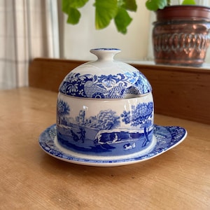 Puede incluir: Un mantequero de cerámica azul y blanco con tapa, con un diseño de paisaje pastoral. El plato se asienta sobre un platillo a juego. El diseño incluye árboles, figuras y edificios, creando una estética clásica y vintage.