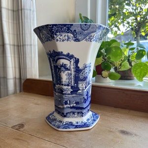 May include: A blue and white ceramic vase with a floral pattern and a scene of a bridge and a building. The vase is octagonal in shape and has a wide mouth.