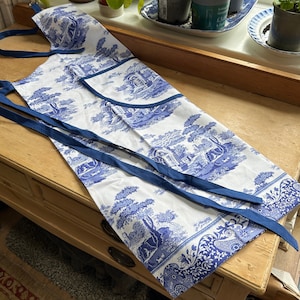 May include: A white apron with a blue and white toile pattern. The apron has two blue straps that tie at the neck and waist.