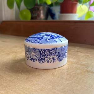 Könnte beinhalten: Kleine, runde Schmuckdose aus Keramik mit Deckel. Die Dose ist weiß mit aufwendigen blauen Blumen- und Paisley-Mustern. Der Deckel zeigt eine detaillierte blau-weiße Landschaftsszene. Ein dünnes Goldband akzentuiert den oberen Rand.