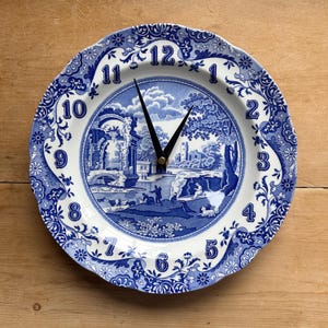 May include: A round wall clock made from a blue and white ceramic plate. The clock face features a pastoral scene with a bridge and figures. Black clock hands indicate the time. The plate has a scalloped edge and decorative floral patterns.