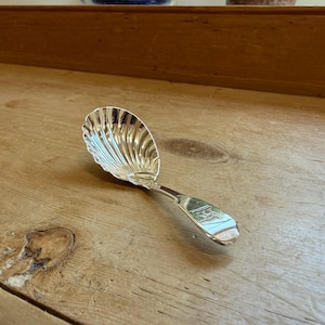 Könnte beinhalten: Ein silberfarbener Löffel mit einer Laffe in Form einer gerippten Muschel. Der Griff ist glatt und flach gestaltet und zeigt in der Mitte ein kleines eingraviertes Wappen oder Emblem mit einer polierten, reflektierenden Metalloberfläche.