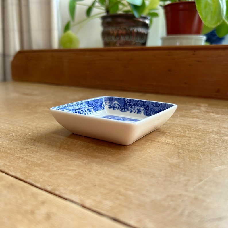 May include: A small, square, white ceramic dish with a blue and white floral pattern. The dish is sitting on a wooden surface.