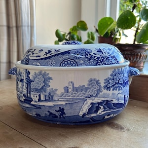 May include: A blue and white ceramic casserole dish with a lid. The dish features a scenic landscape with a castle and trees. The lid has a floral pattern.