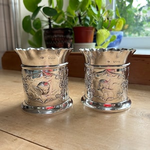 May include: Two silver, ornate, decorative cups with a scalloped rim. Each cup features an embossed design of an angel with wings and a floral border. The cups are on a wooden surface.